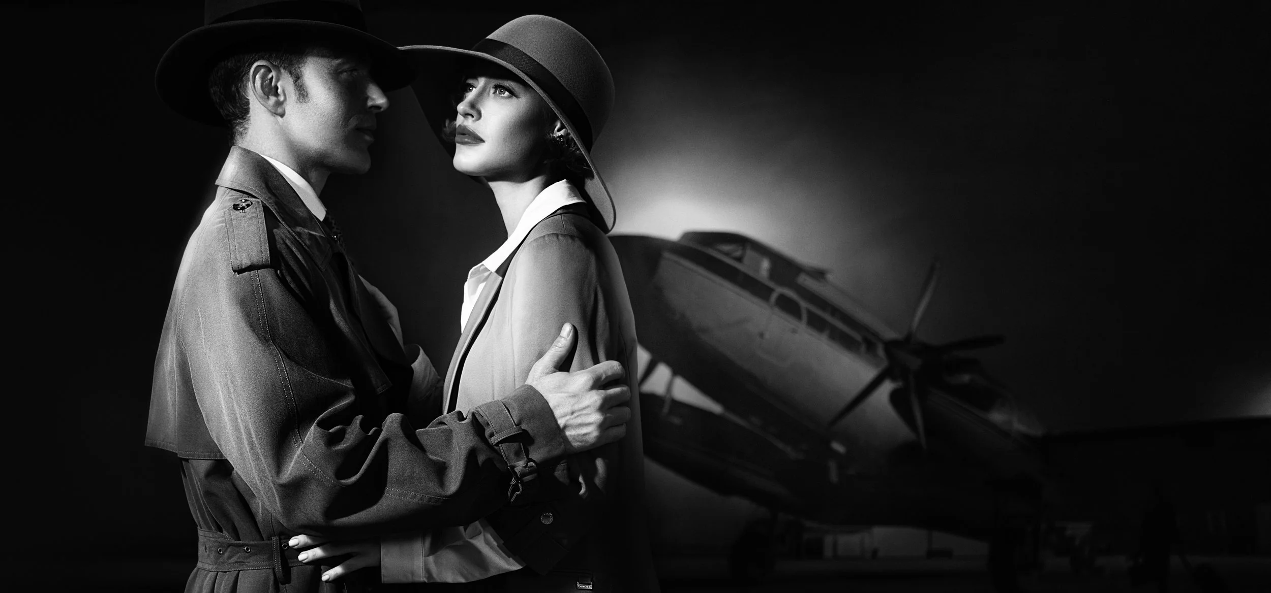 Black and white cinematic portrait of a couple in period dress beside a vintage aircraft